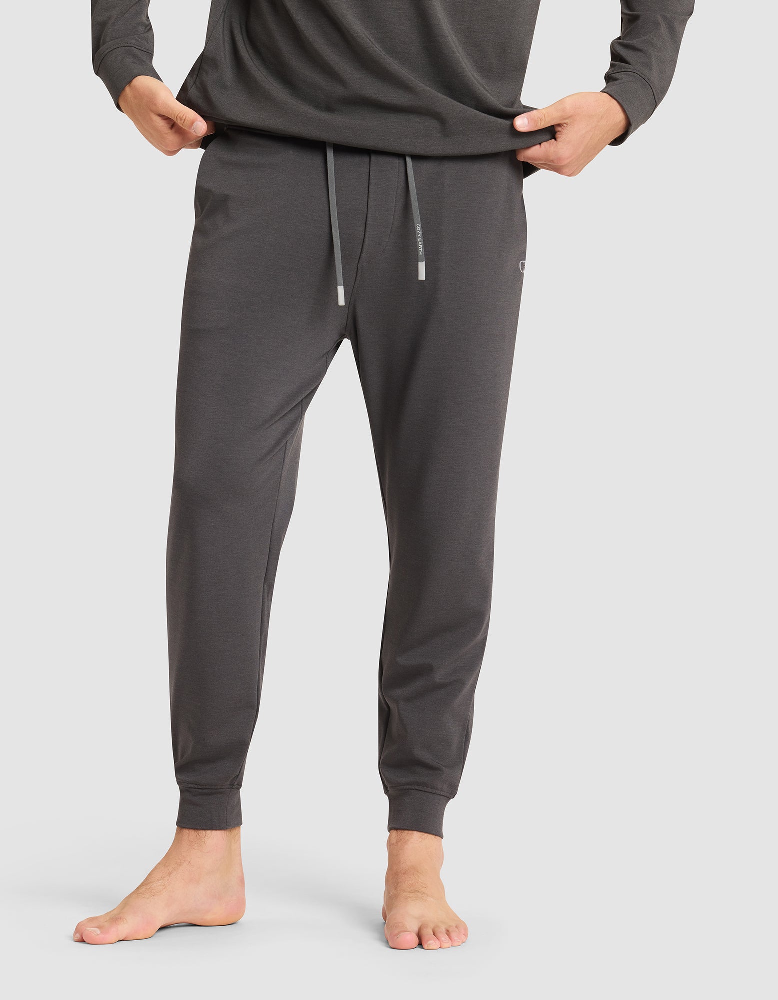 A man stands barefoot against a plain light background, wearing Cozy Earth's Men's Brushed Bamboo Jogger Pant in dark gray with hands in pockets, paired with a matching long-sleeve shirt. 