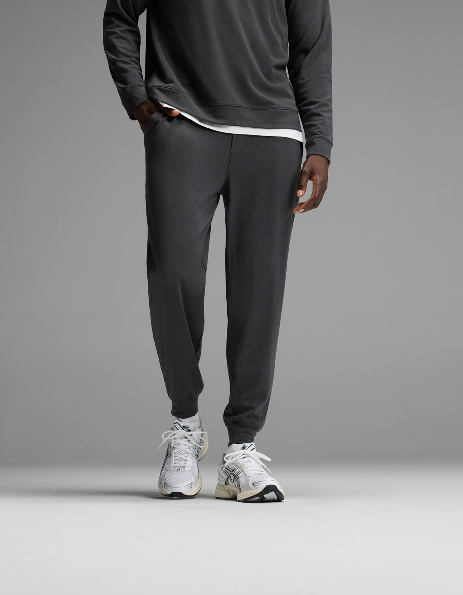 A person stands against a plain gray background wearing Cozy Earth’s Men’s Brushed Bamboo Joggers in Charcoal, paired with a matching long-sleeve top layered over a white undershirt and finished with white athletic sneakers. The upper body is partially out of frame.