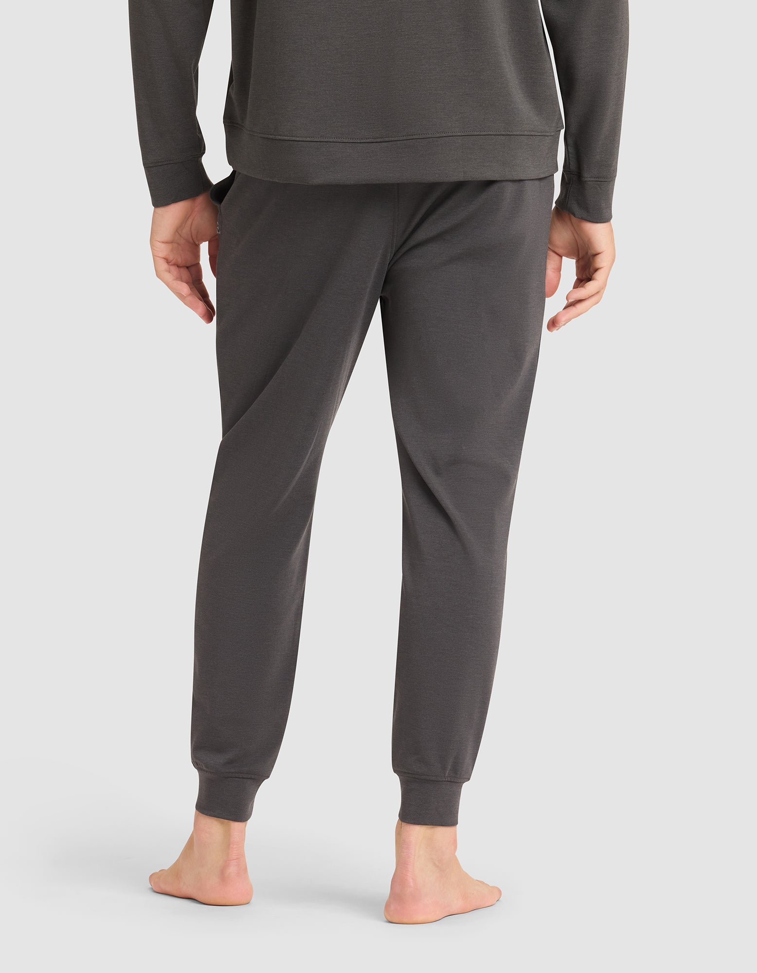 A person stands barefoot on a white background, shown from the back wearing Cozy Earth's Men's Brushed Bamboo Jogger Pant in dark gray paired with a matching long-sleeve top. 
