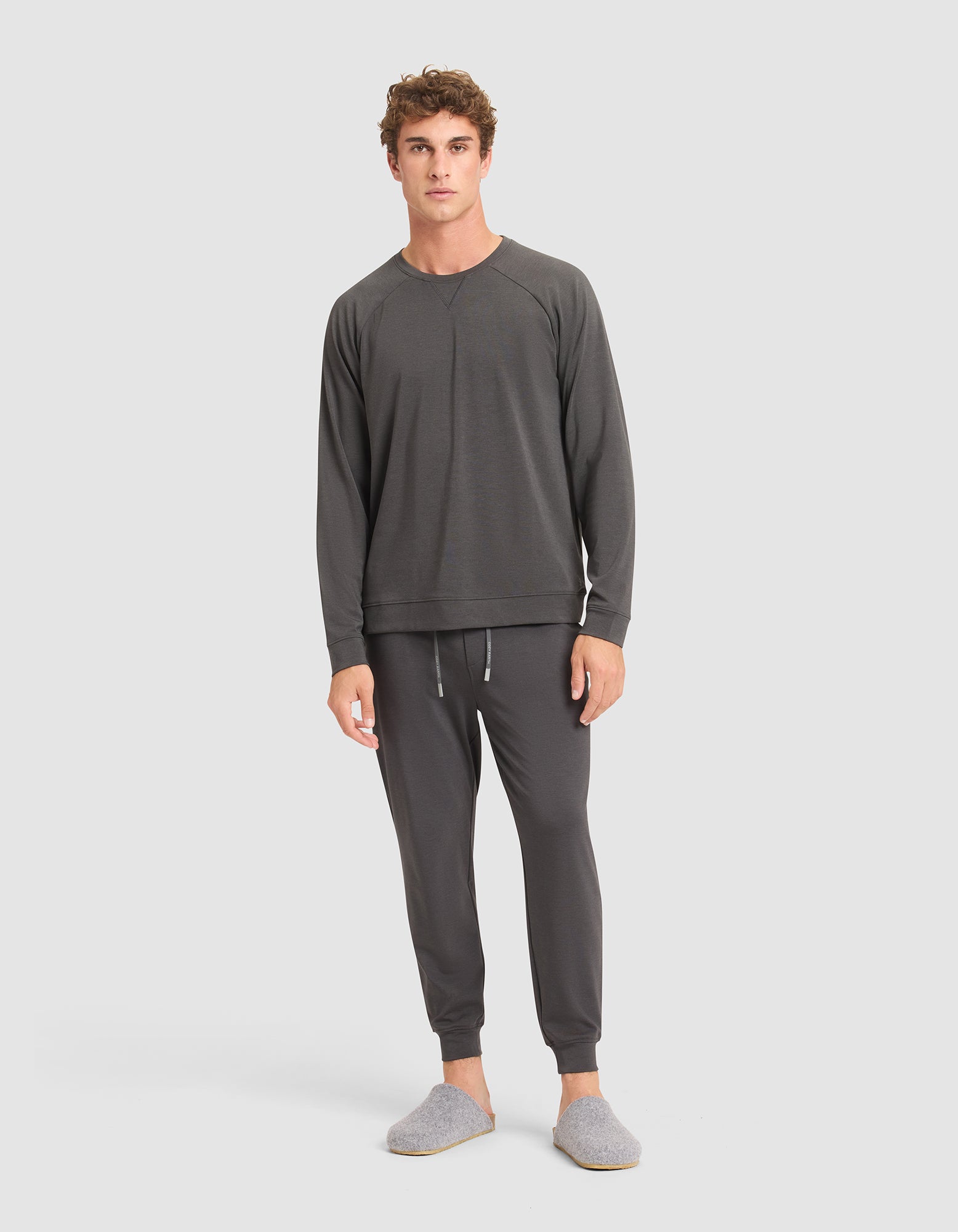 A young man wearing Cozy Earth's Men's Brushed Bamboo Jogger Pant in dark gray, paired with a matching long-sleeve top and gray slippers, stands against a plain white background, looking at the camera. 