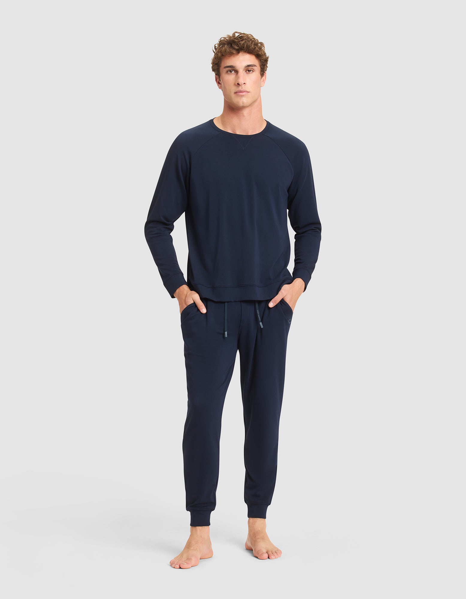 A young man stands barefoot against a light gray background, wearing Cozy Earth's Men's Brushed Bamboo Jogger Pant in navy blue with a matching long-sleeve sweatshirt and his hands in his pockets.