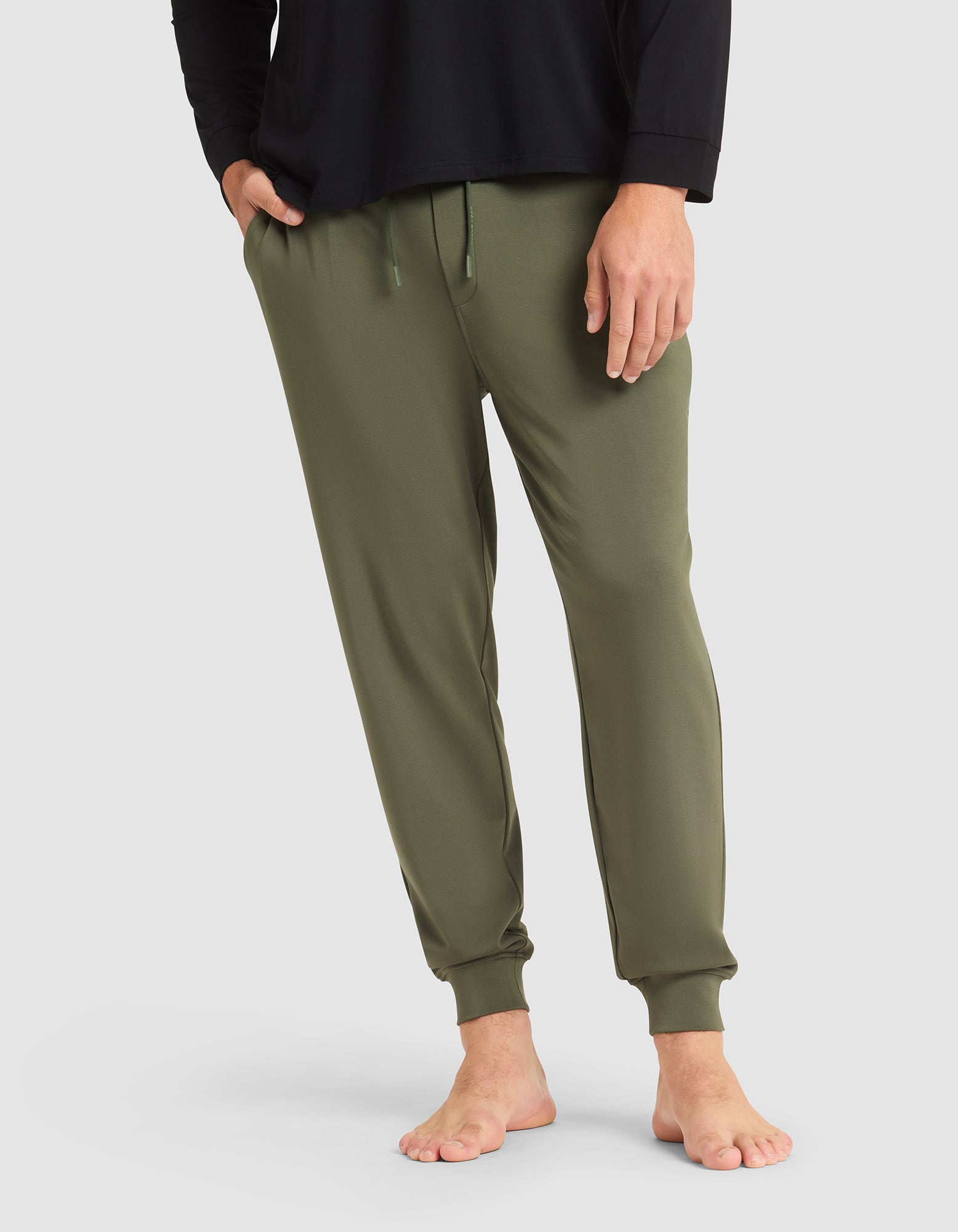 A person models the Cozy Earth Men's Brushed Bamboo Jogger Pant in olive green, shown barefoot with a black long-sleeve shirt against a plain light gray background. Only the lower half of the body is visible. 