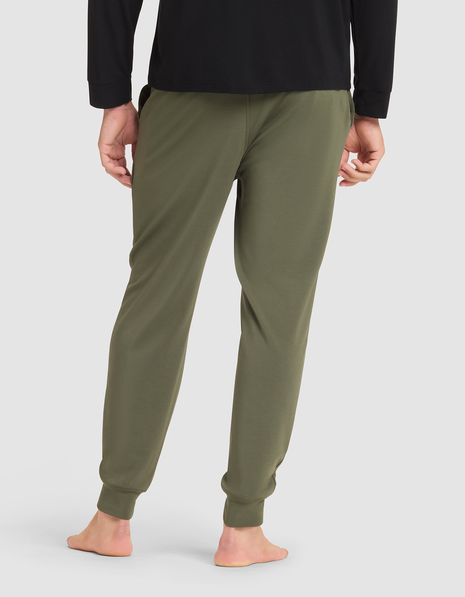 A person is standing barefoot, viewed from the back against a plain light background, wearing Cozy Earth's Men's Brushed Bamboo Jogger Pant in olive green and a black long-sleeve shirt. 