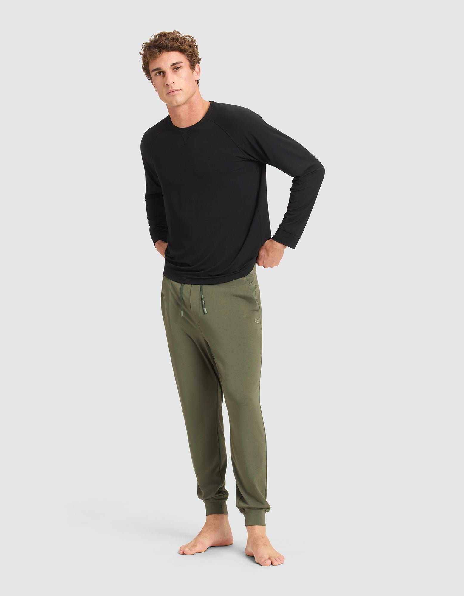 A man stands barefoot against a plain background, wearing a long-sleeve black shirt and Cozy Earth Men's Brushed Bamboo Jogger Pant in olive green, with his hands in his pockets. 