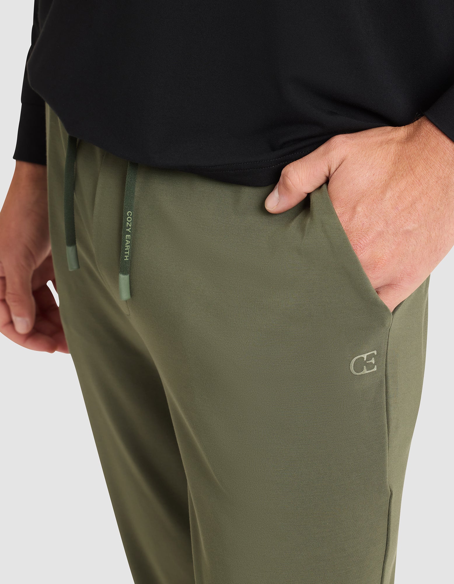 A man wears Cozy Earth Men's Brushed Bamboo Jogger Pant in olive green, featuring a drawstring waistband and logo, shown from the waist down with one hand in the pocket, paired with a black long-sleeve shirt. 