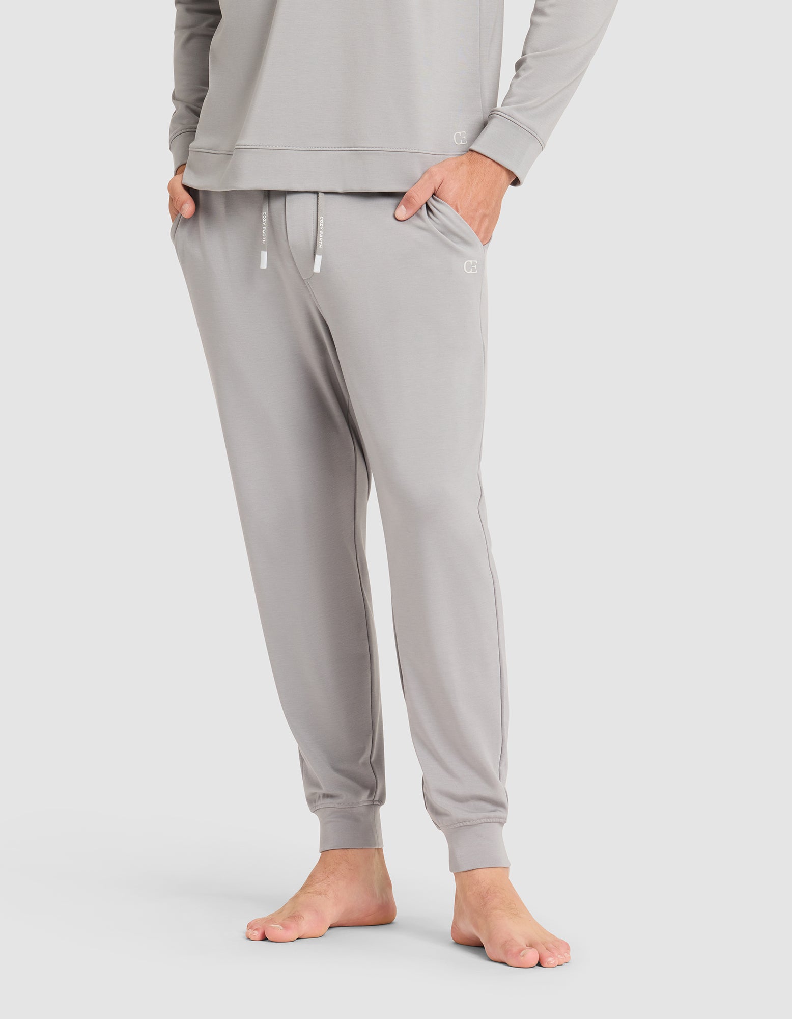 A person stands barefoot against a plain light background, wearing Cozy Earth's Men's Brushed Bamboo Jogger Pant in light gray with hands in pockets, paired with a matching long-sleeve top. 