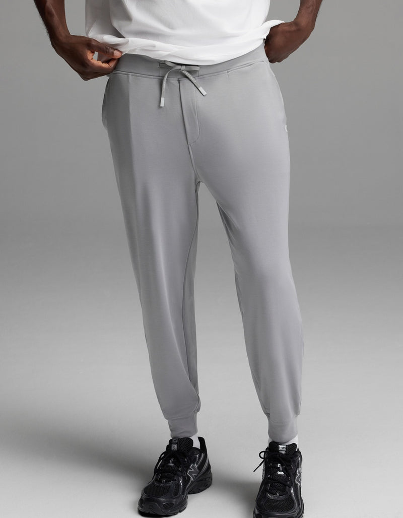 A person is shown from the shoulders down, wearing Cozy Earth’s Men’s Brushed Bamboo Joggers in light gray, paired with a white shirt and black athletic shoes, standing against a plain gray background.