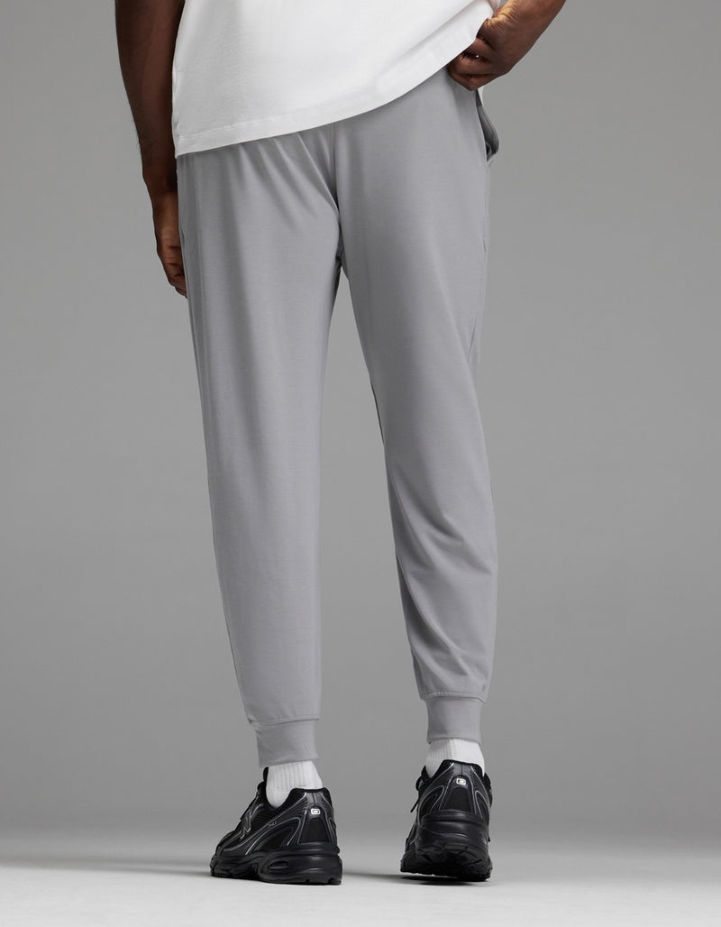 A person is shown from the back, standing against a plain gray background, wearing Cozy Earth Men’s Brushed Bamboo Joggers in light gray paired with a white shirt and black sneakers.