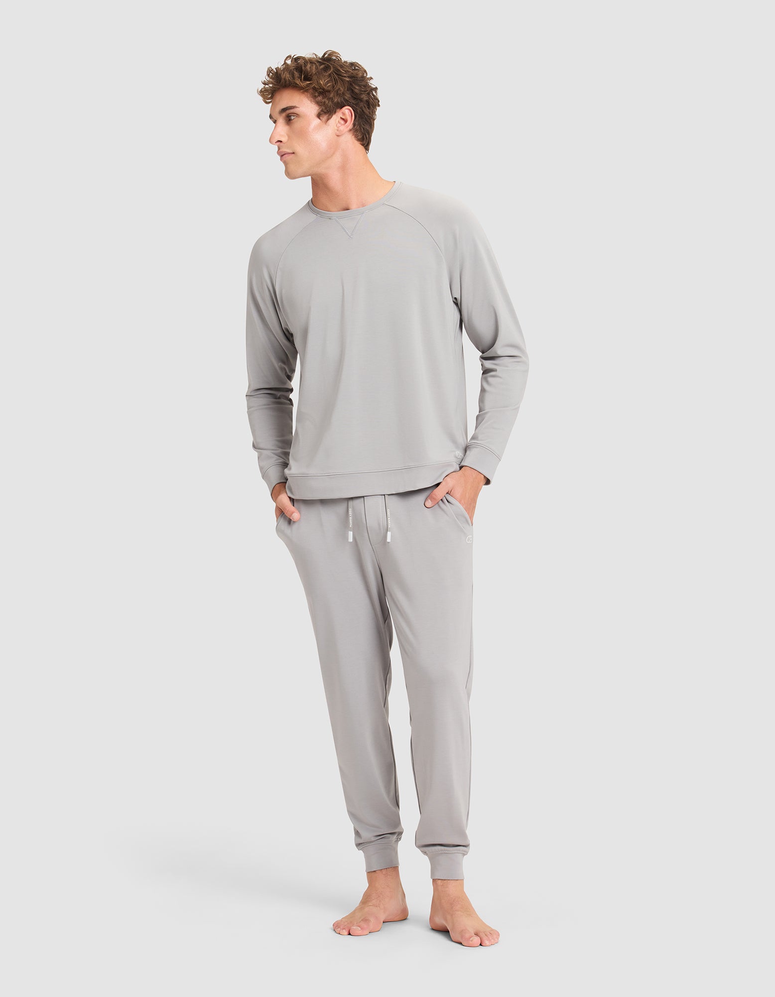 A man stands barefoot against a plain background, wearing the Cozy Earth Men's Brushed Bamboo Jogger in light gray, with his hands in his pockets and looking slightly to the side. 