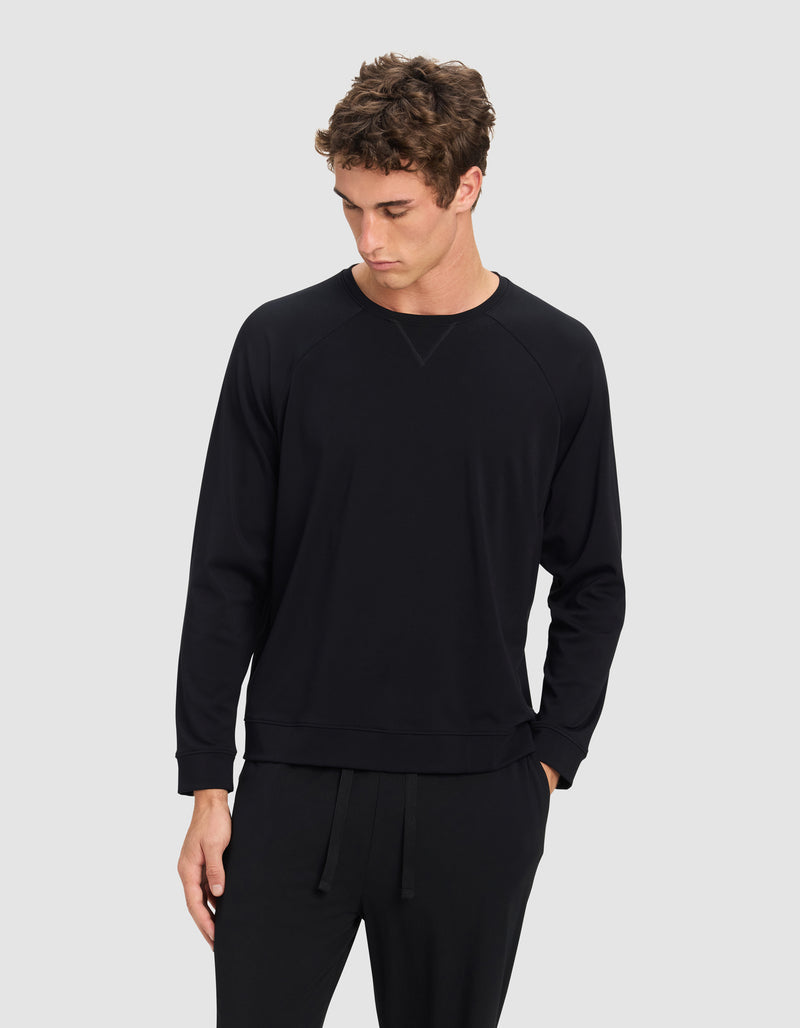 A young man with curly brown hair wears the Cozy Earth Men's Brushed Bamboo Pullover Crew in black, paired with black pants, standing against a plain light grey background and looking down to his left.