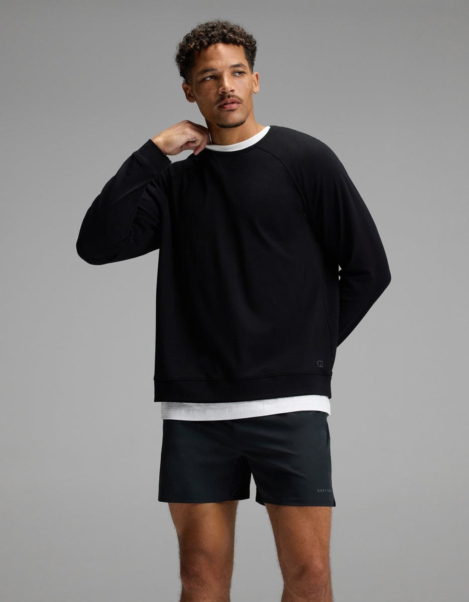 A man stands against a gray background, wearing the Cozy Earth Men’s Brushed Bamboo Pullover Crew in black over a white shirt and black shorts. He has short curly hair and is touching his neck with his right hand, looking slightly to the side.|Color:Black
