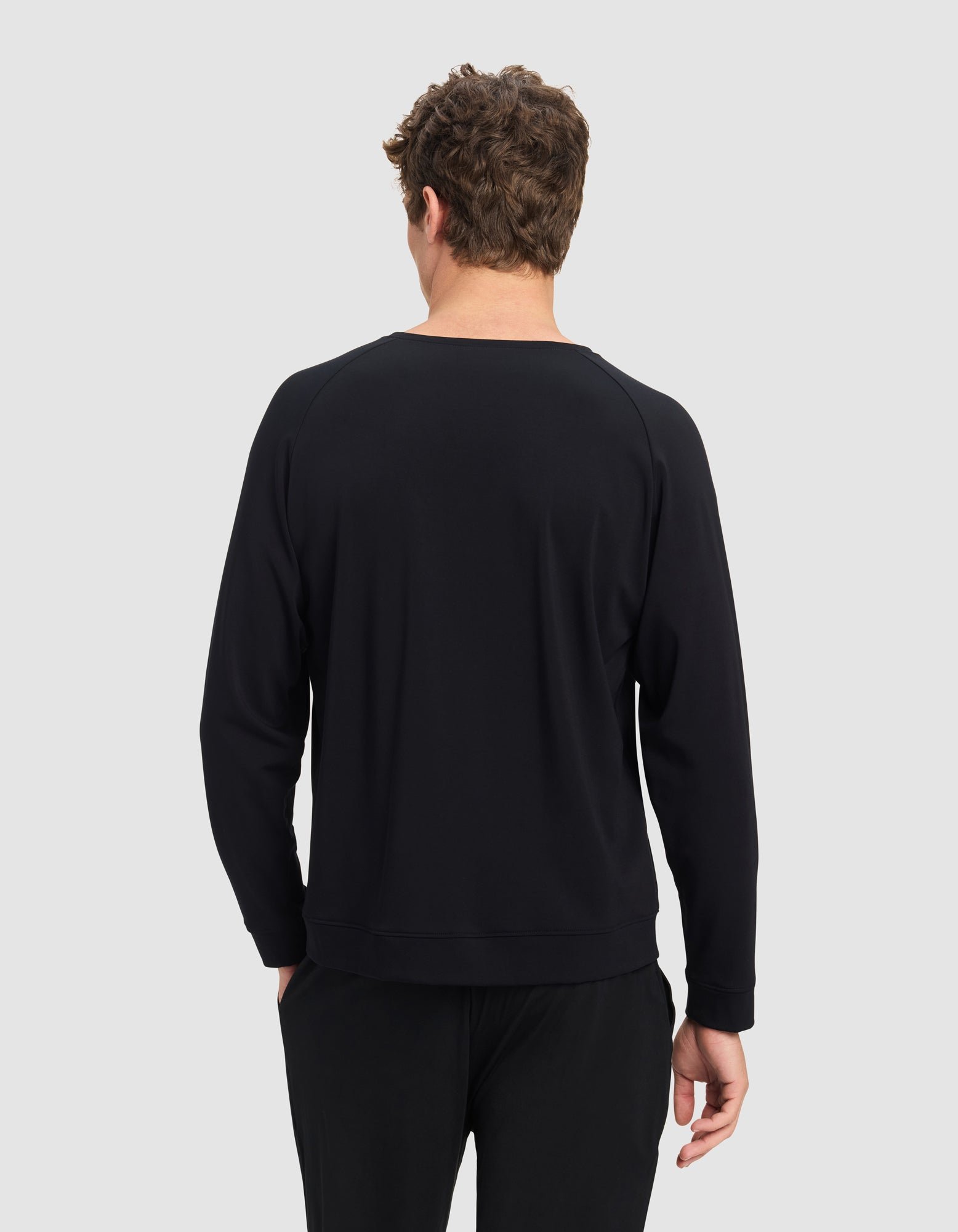 A man with short curly hair stands with his back to the camera, wearing a plain black Cozy Earth Men's Brushed Bamboo Pullover Crew and black pants against a light gray background.