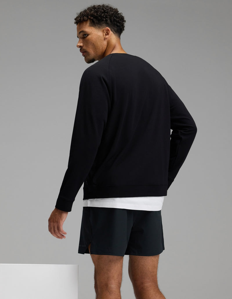 A man with short curly hair is wearing the Cozy Earth Men’s Brushed Bamboo Pullover Crew in black over a white shirt and paired with black shorts, standing and looking over his shoulder against a plain gray background.