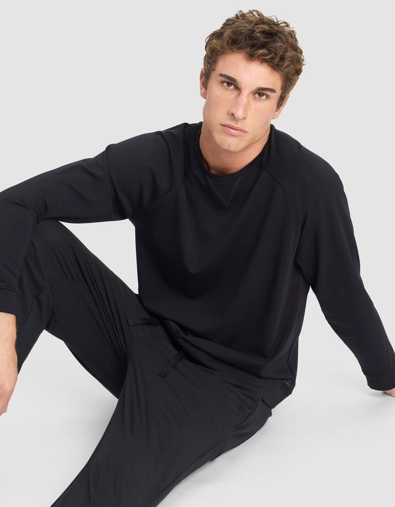 A young man with short curly hair wears the Cozy Earth Men's Brushed Bamboo Pullover Crew and matching pants, sitting on the floor against a light gray background and looking at the camera with a neutral expression.