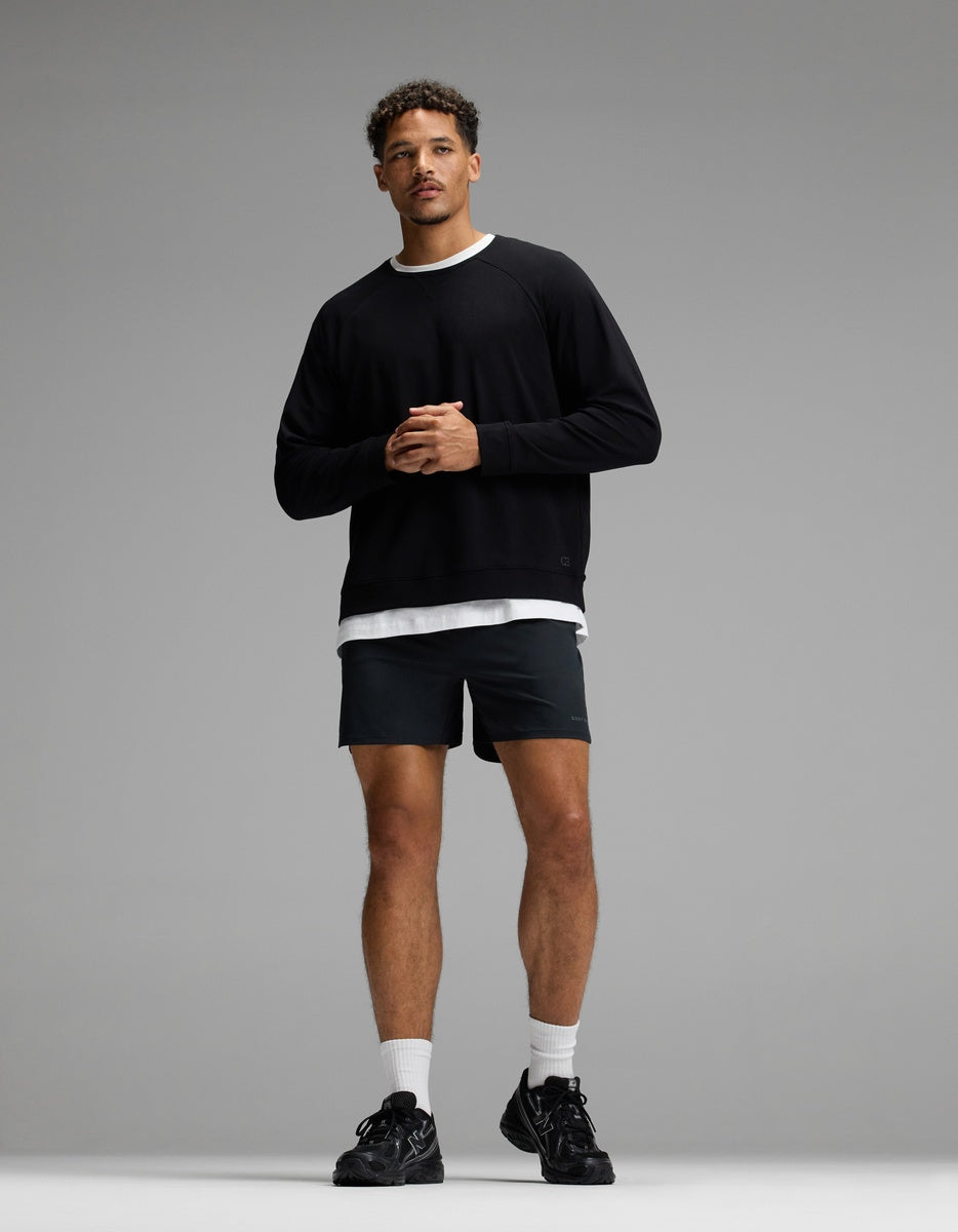 A man stands against a gray background wearing the Cozy Earth Men’s Brushed Bamboo Pullover Crew, paired with black shorts, white crew socks, and black sneakers. He has short curly hair and his hands are clasped in front of him.