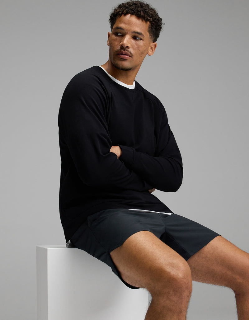 A man with short curly hair sits on a white block, wearing Cozy Earth’s Men’s Brushed Bamboo Pullover Crew in black over a white shirt and dark shorts, looking to his left against a plain gray background.
