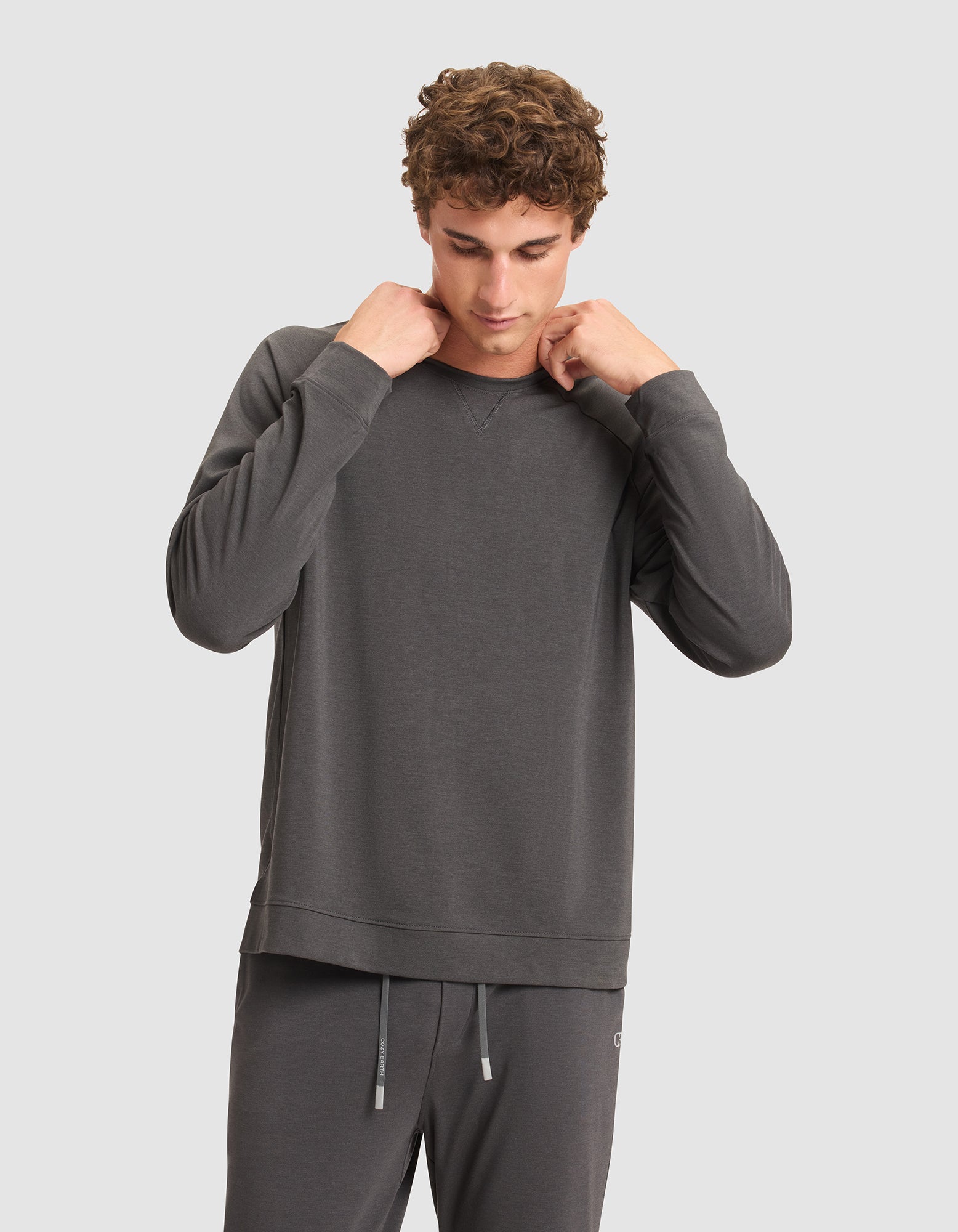 A man with curly hair adjusts the collar of his Cozy Earth Men's Brushed Bamboo Pullover Crew, paired with matching gray pants, while standing against a plain light gray background. 
