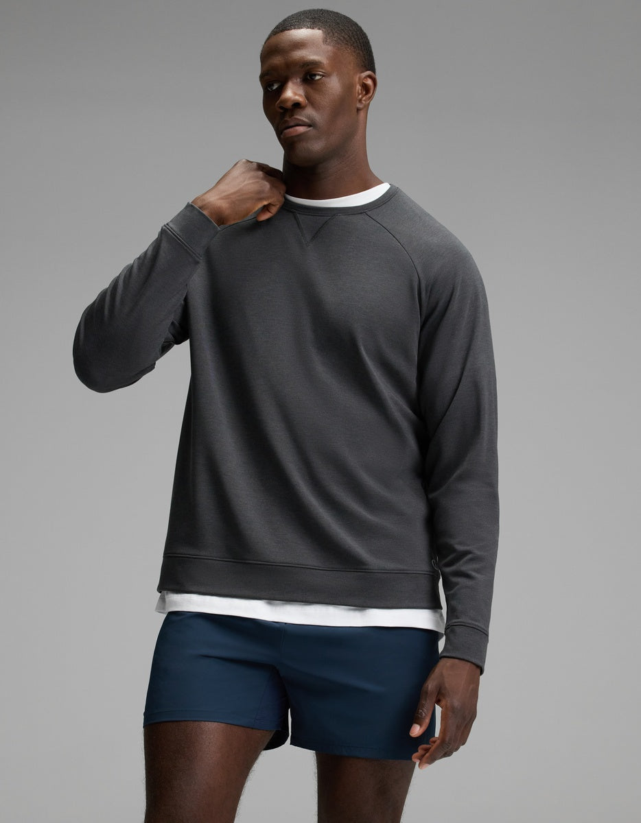 A man wearing the Cozy Earth Men’s Brushed Bamboo Pullover Crew in Charcoal over a white shirt and navy blue shorts stands against a plain gray background, gazing slightly to the side with one hand near his neck.