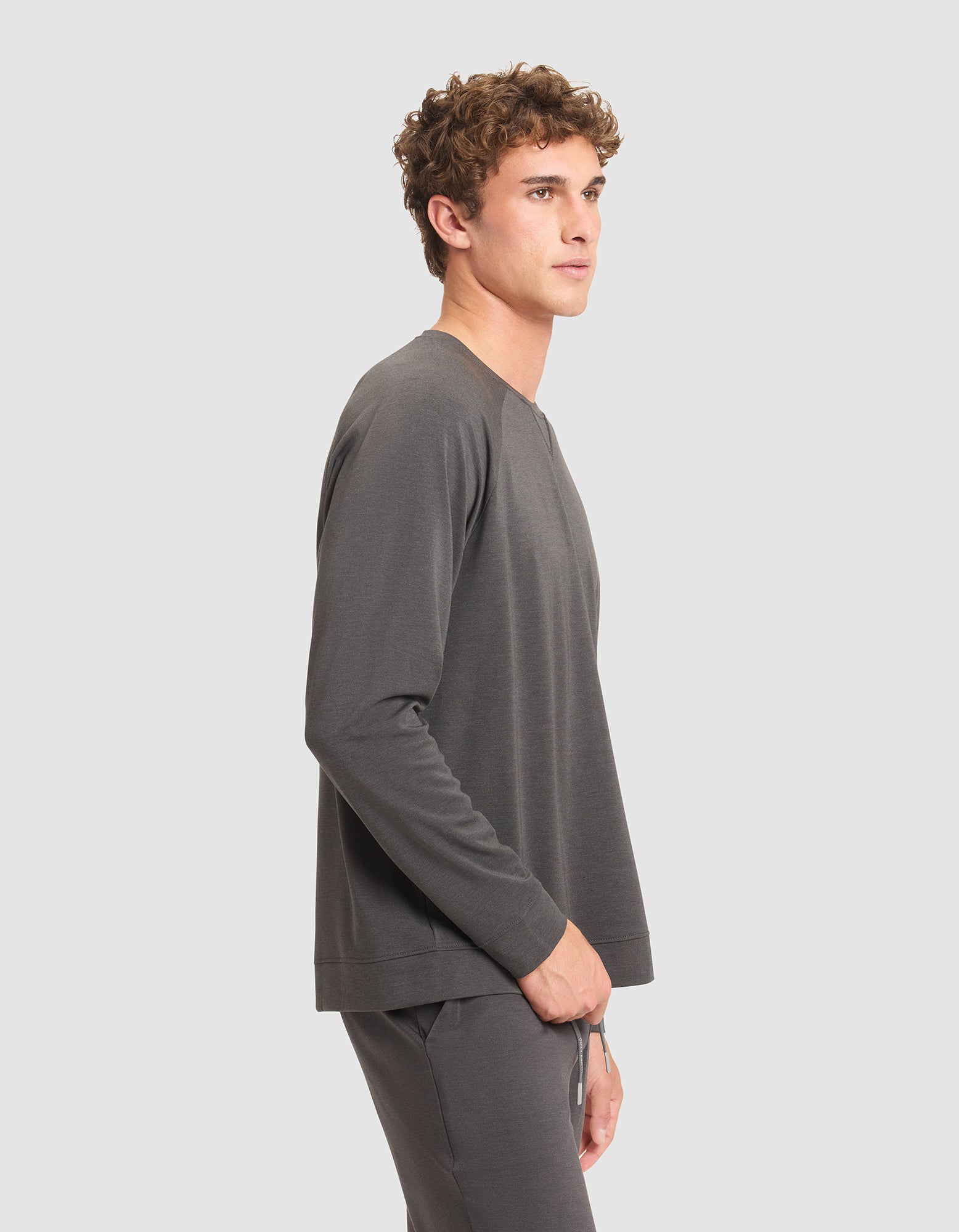 A young man with curly hair stands in profile against a plain light gray background, wearing a matching dark gray Men's Brushed Bamboo Pullover Crew by Cozy Earth.  