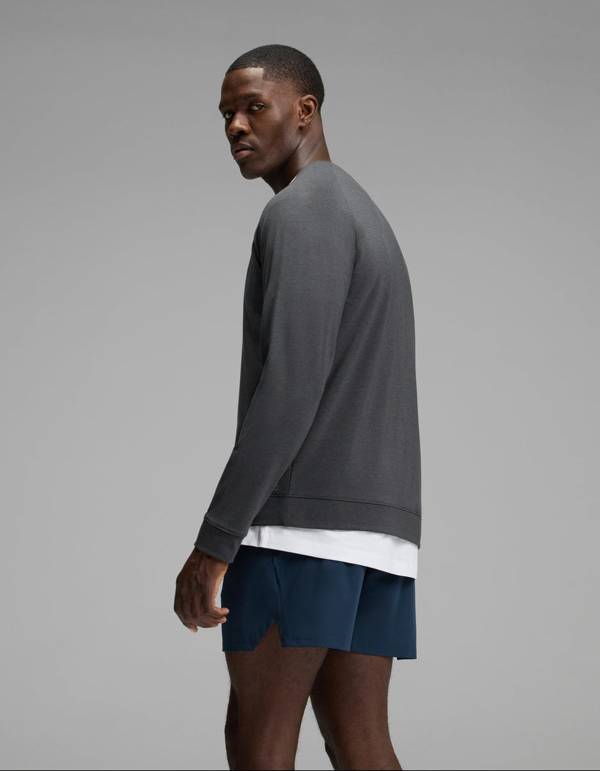A man wearing the Cozy Earth Men’s Brushed Bamboo Pullover Crew in gray, layered over a white undershirt and paired with blue shorts, stands sideways while turning his head toward the camera against a plain gray background.