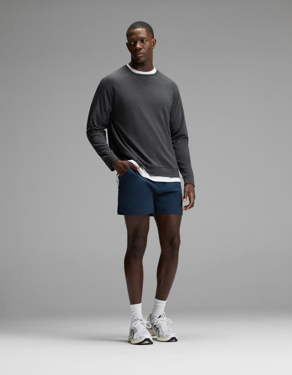 A man stands against a plain gray background, wearing the Cozy Earth Men’s Brushed Bamboo Pullover Crew in gray, layered over a white shirt, paired with navy shorts, white crew socks, and white athletic shoes.