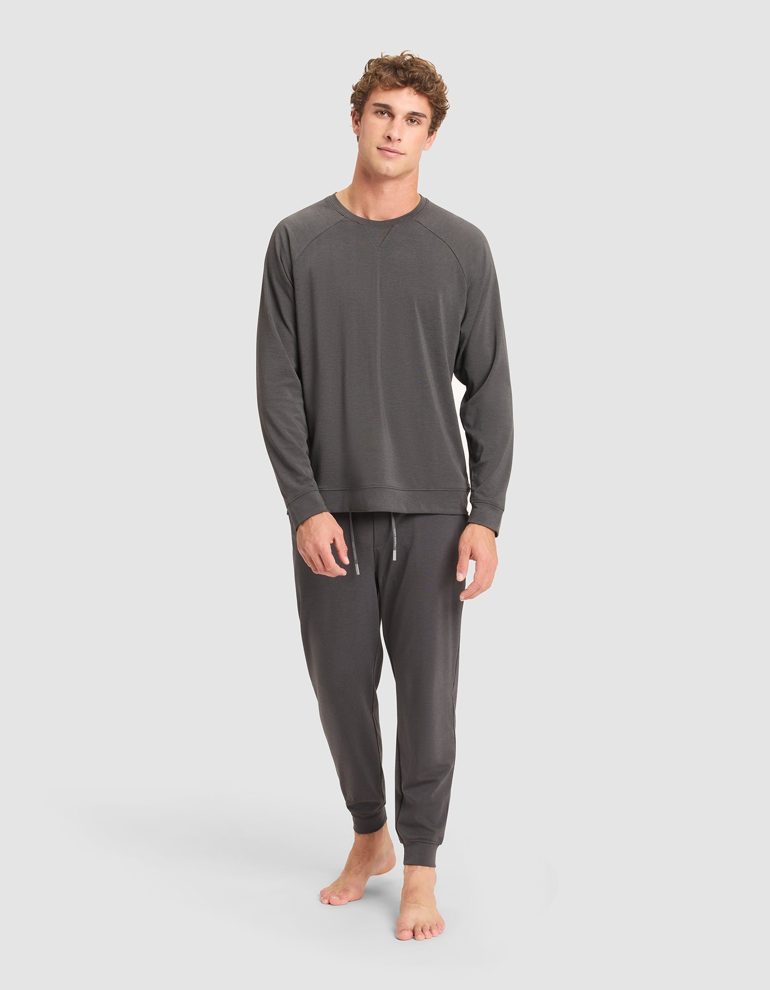 A young man stands barefoot against a plain background, wearing the Cozy Earth Men's Brushed Bamboo Pullover Crew in dark gray lounge set. He appears relaxed, with one hand by his side and the other hanging loosely.  