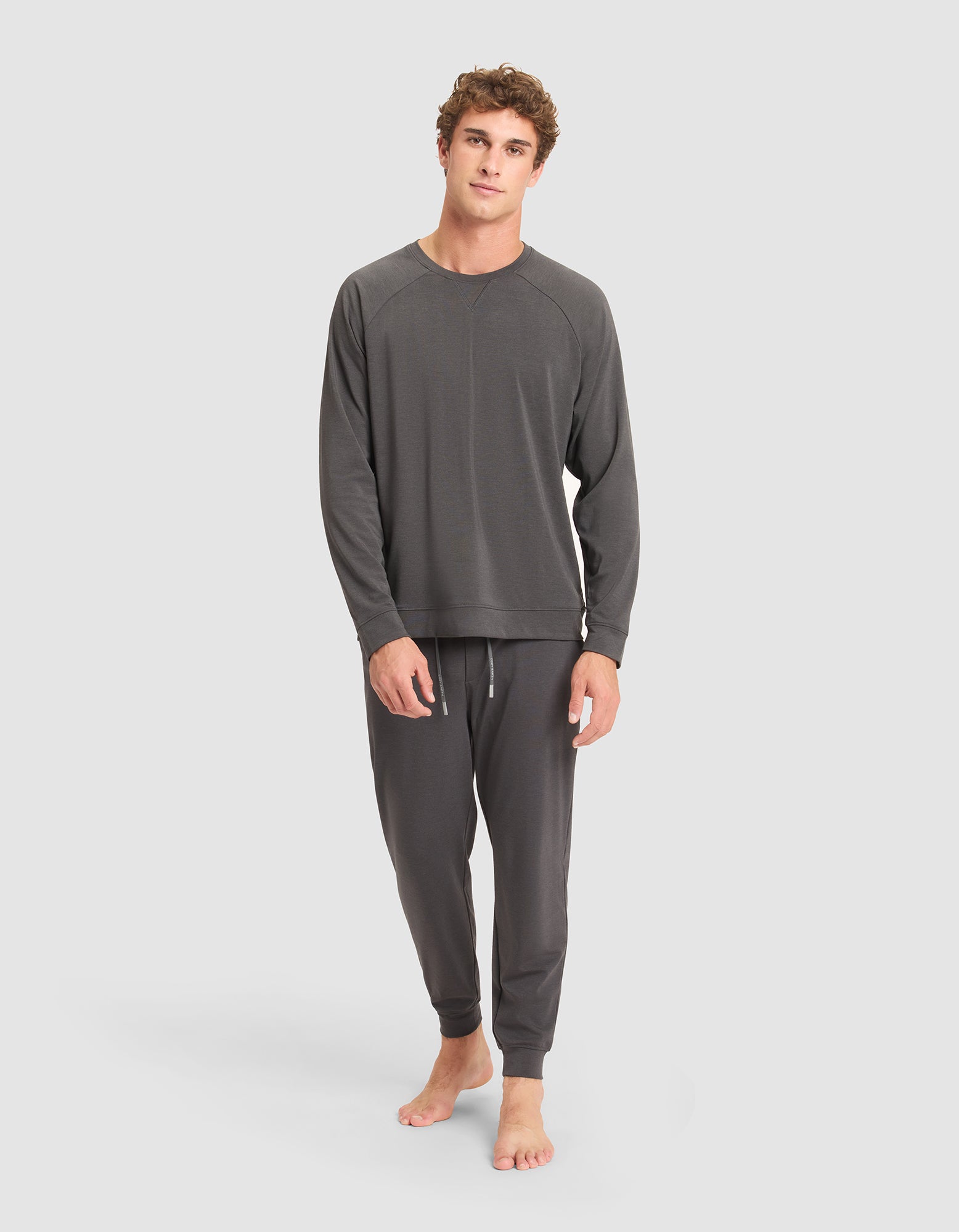 A young man stands barefoot against a plain background, wearing the Cozy Earth Men's Brushed Bamboo Pullover Crew in dark gray lounge set. He appears relaxed, with one hand by his side and the other hanging loosely.  