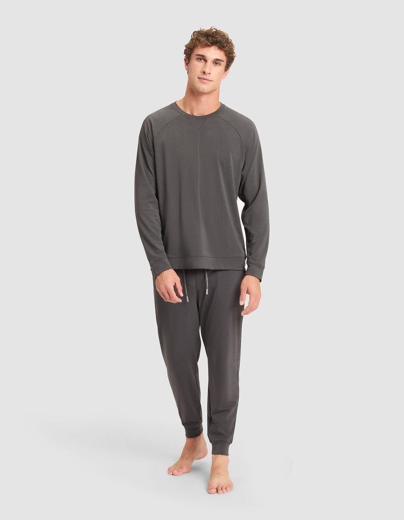 A young man stands barefoot against a plain background, wearing the Cozy Earth Men's Brushed Bamboo Pullover Crew in dark gray lounge set. He appears relaxed, with one hand by his side and the other hanging loosely.