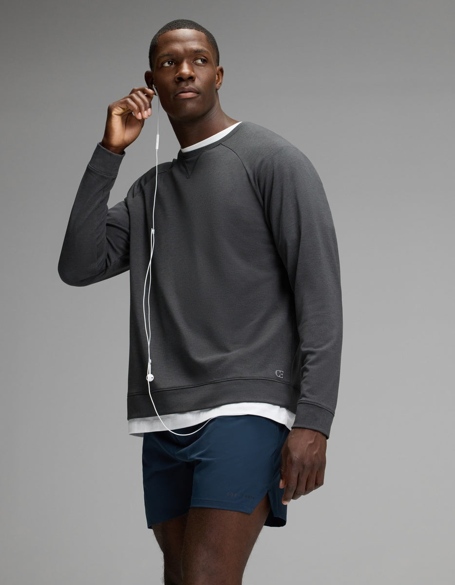 A man wearing the Cozy Earth Men’s Brushed Bamboo Pullover Crew in dark grey over a white shirt and paired with navy shorts stands against a plain background, holding one earbud to his ear with a relaxed expression.