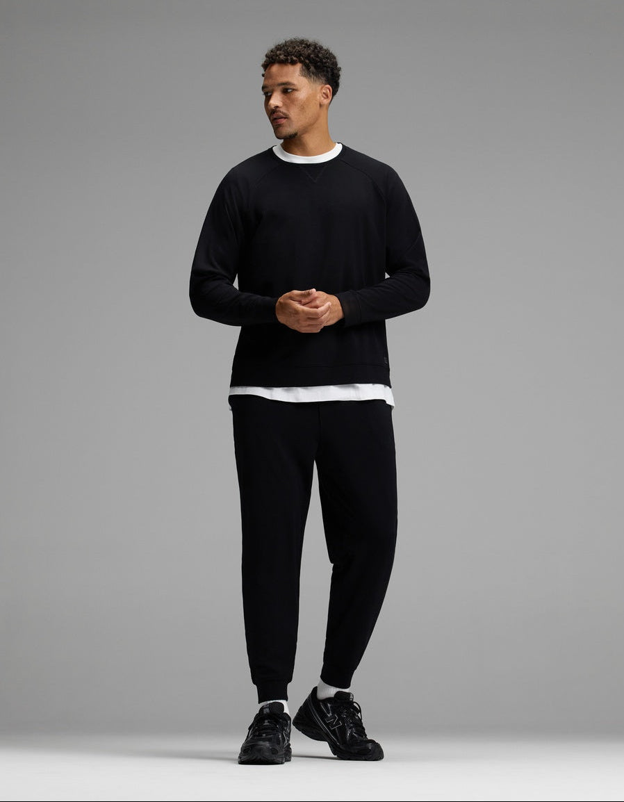 A man stands in a studio wearing the Cozy Earth Men’s Brushed Bamboo Jogger Set in black, paired with black sneakers and a white undershirt. He is looking to the side with his hands clasped.|Color:Black