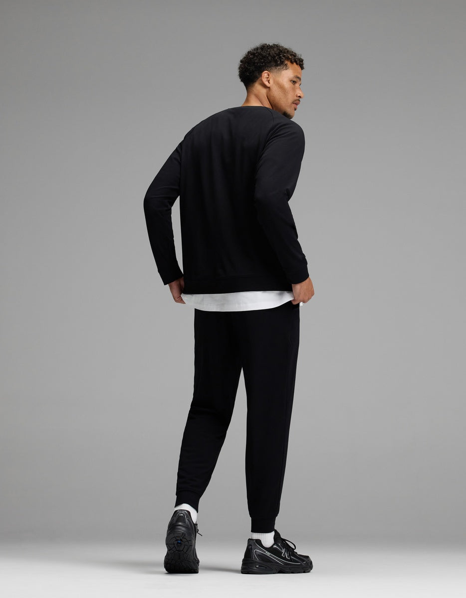 A man stands against a gray background, facing away and looking over his shoulder. He wears the Men’s Brushed Bamboo Jogger Set by Cozy Earth, featuring a black long-sleeve top and matching black joggers layered over a white shirt, paired with white socks and black sneakers.