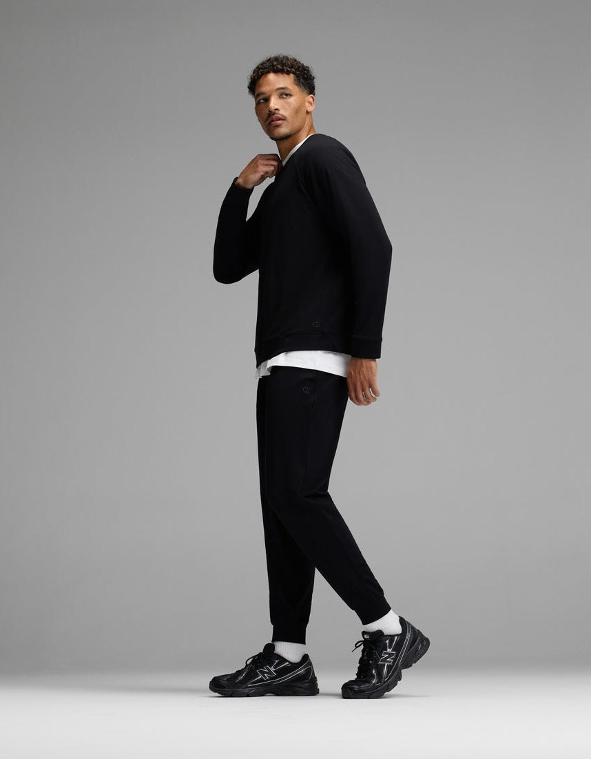 A man stands in a studio wearing the Cozy Earth Men’s Brushed Bamboo Jogger Set in black, paired with black sneakers and a white undershirt. He looks to the side with one hand near his neck, against a plain gray background.