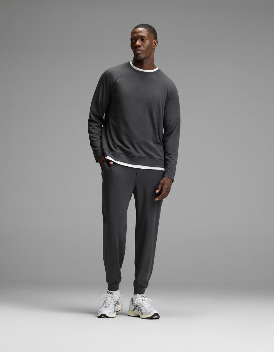 A man stands against a plain gray background, wearing Cozy Earth’s Men’s Brushed Bamboo Jogger Set in dark gray over a white layered shirt and white sneakers, with his hands in his pockets.