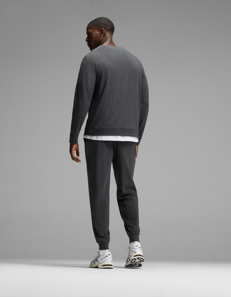 A man stands facing away, wearing the Cozy Earth Men’s Brushed Bamboo Jogger Set in dark gray over a white shirt, paired with white athletic shoes, against a plain gray background.