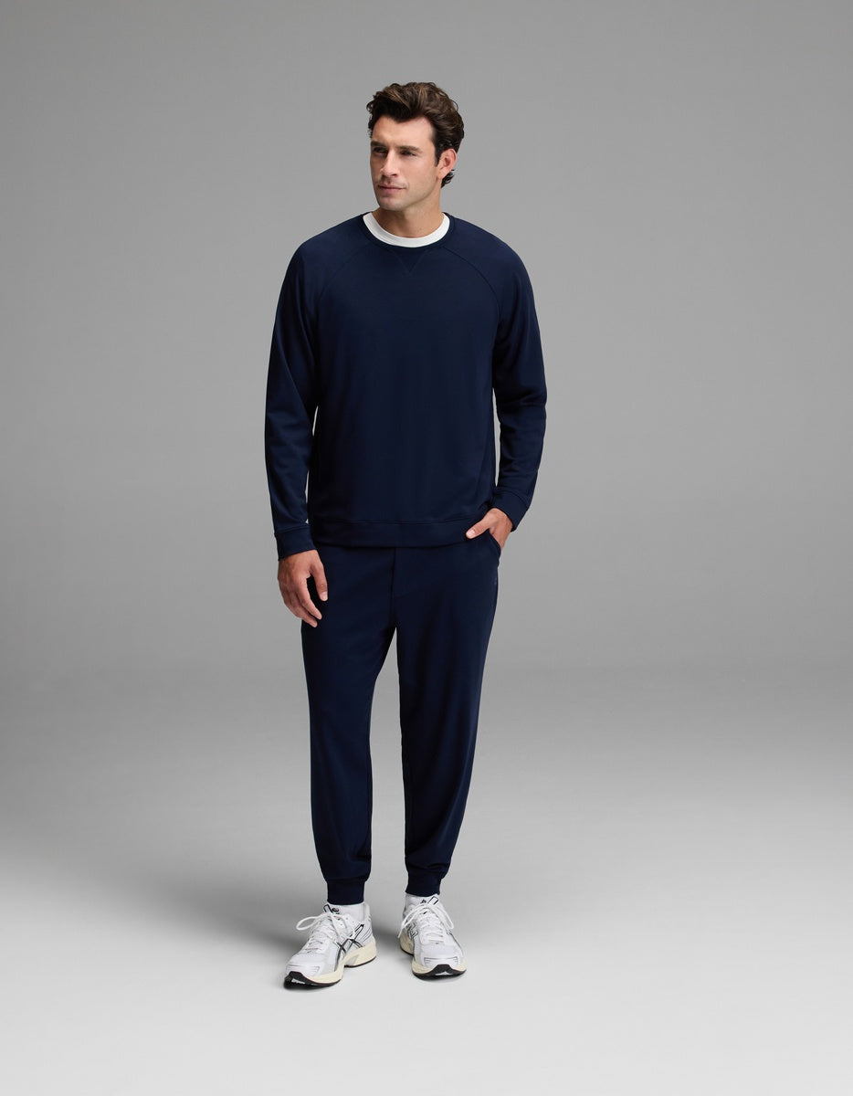 A man is seen standing against a plain gray background wearing the Cozy Earth Men’s Brushed Bamboo Jogger Set in navy blue, paired with white sneakers and a white shirt underneath. He looks slightly to his left with one hand in his pocket.|Color:Navy