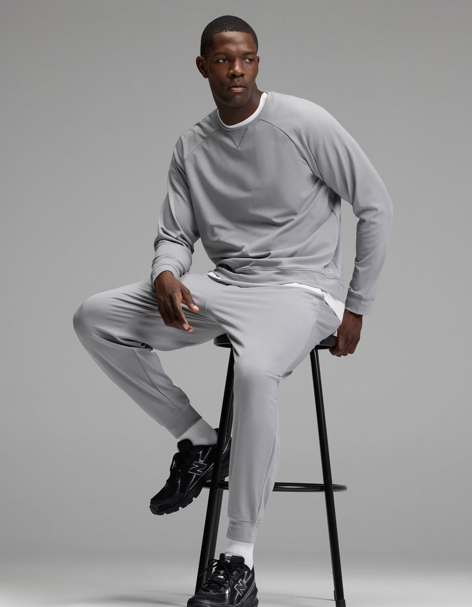A man wearing the Cozy Earth Men’s Brushed Bamboo Jogger Set in light gray sits on a tall black stool against a plain gray background. He has short hair and is wearing black athletic shoes.|Color:Stone