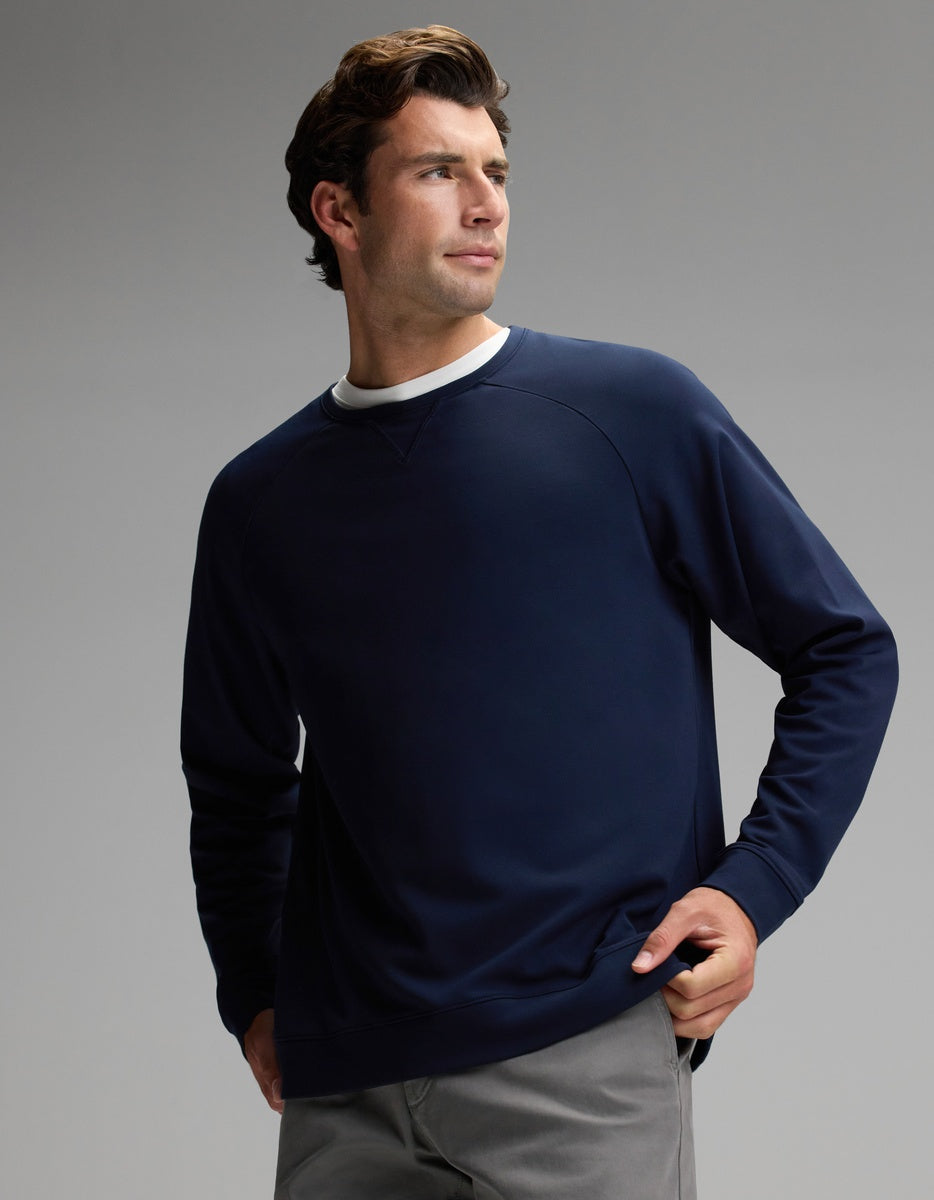 A man wears the Cozy Earth Men’s Brushed Bamboo Pullover Crew in navy, paired with gray pants, as he stands against a plain gray background and looks off to the side with one hand in his pocket.