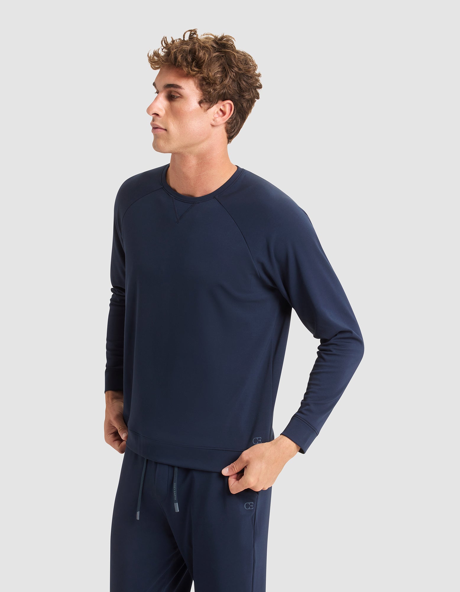 A young man with curly hair, dressed in Cozy Earth's Men's Brushed Bamboo Pullover Crew, stands against a plain light gray background, gazing thoughtfully to the side. 