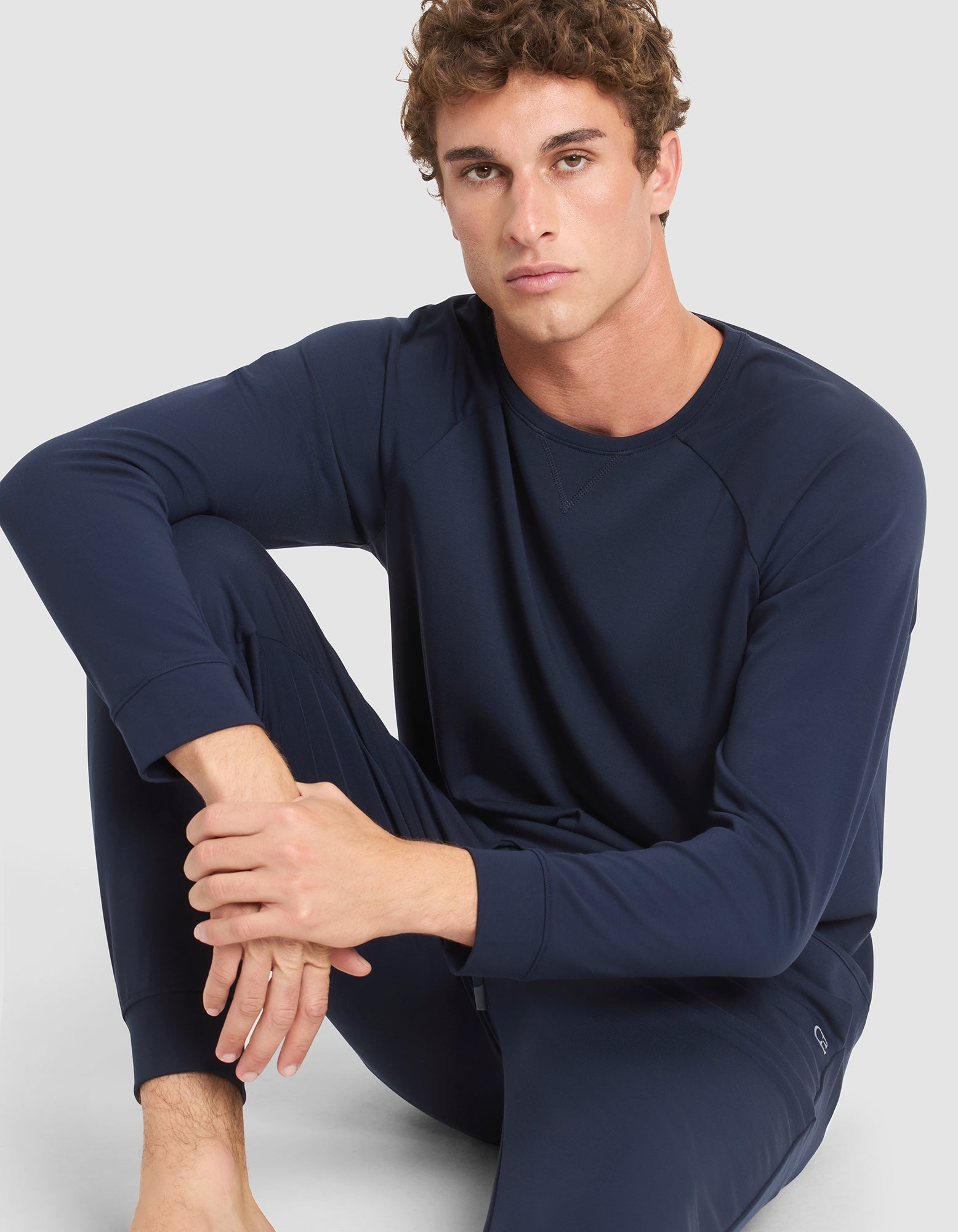 A young man with curly hair wears the Cozy Earth Men's Brushed Bamboo Pullover Crew in navy blue, sitting barefoot on the floor with one arm resting on his knee against a plain light background. 