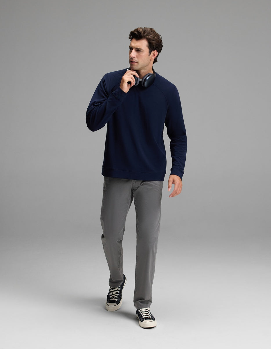 A man wearing the Cozy Earth Men’s Brushed Bamboo Pullover Crew in navy, paired with gray pants and black sneakers, walks forward holding black headphones near his chin against a plain light gray background.