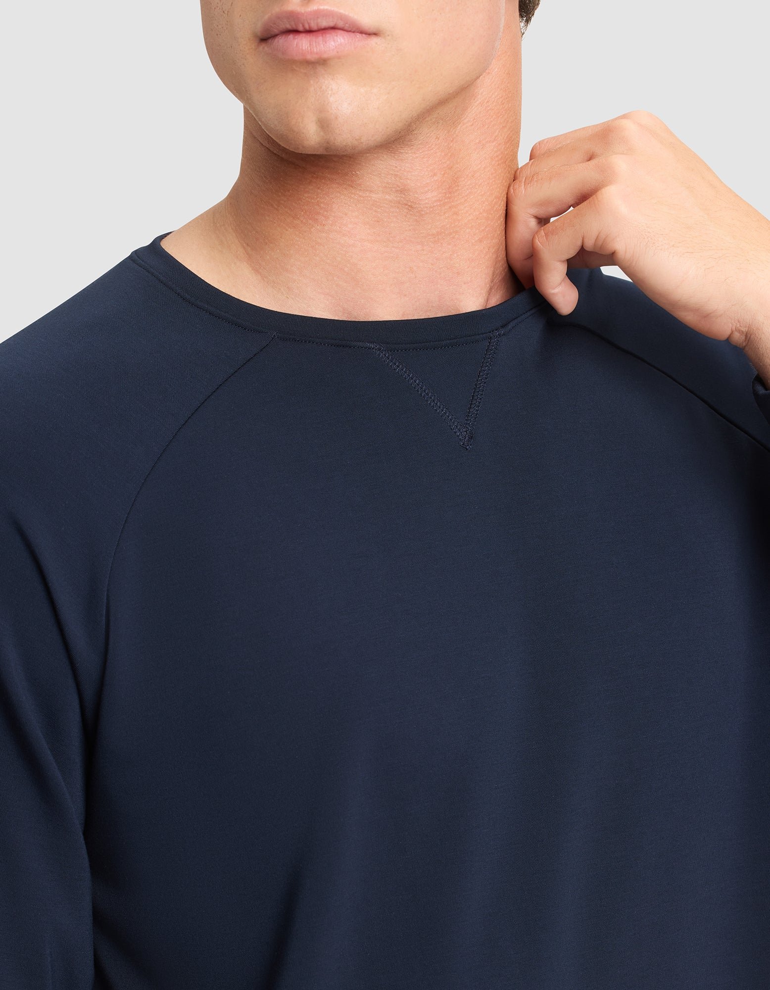 A man models the Cozy Earth Men's Brushed Bamboo Pullover Crew in navy blue, with only his lower face and upper torso visible as he stands against a light gray background, touching his neck with his right hand. 