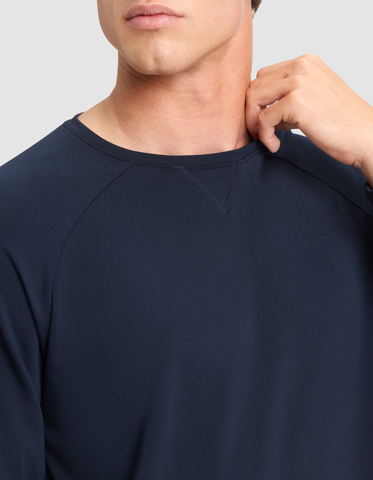A man models the Cozy Earth Men's Brushed Bamboo Pullover Crew in navy blue, with only his lower face and upper torso visible as he stands against a light gray background, touching his neck with his right hand. 