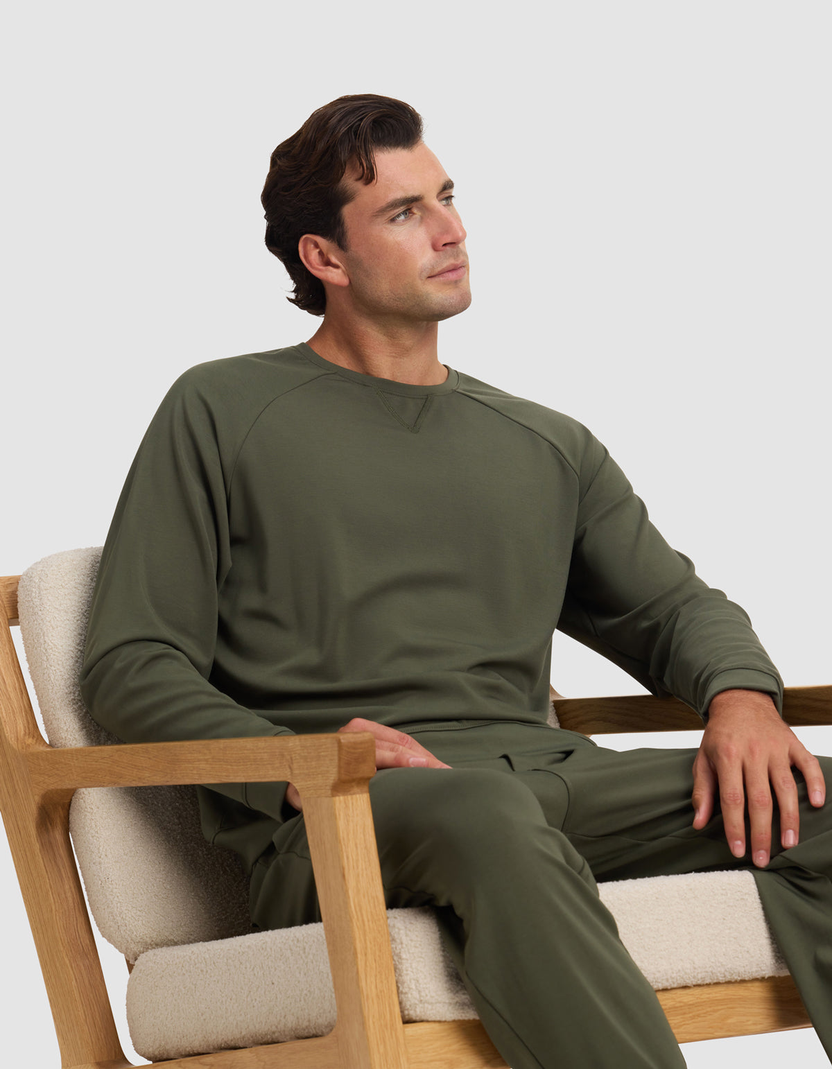 A man wearing the Cozy Earth Men's Brushed Bamboo Pullover Crew in olive sits calmly in a light-cushioned wooden chair, gazing upward against a plain background.