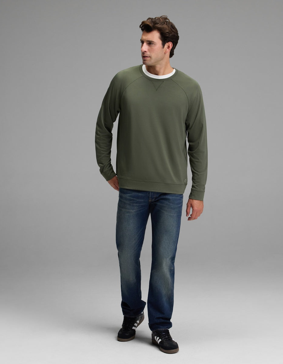 A man wearing the Cozy Earth Men’s Brushed Bamboo Pullover Crew in green, paired with blue jeans and black sneakers, stands against a plain gray background, looking to his left with one hand in his pocket.
