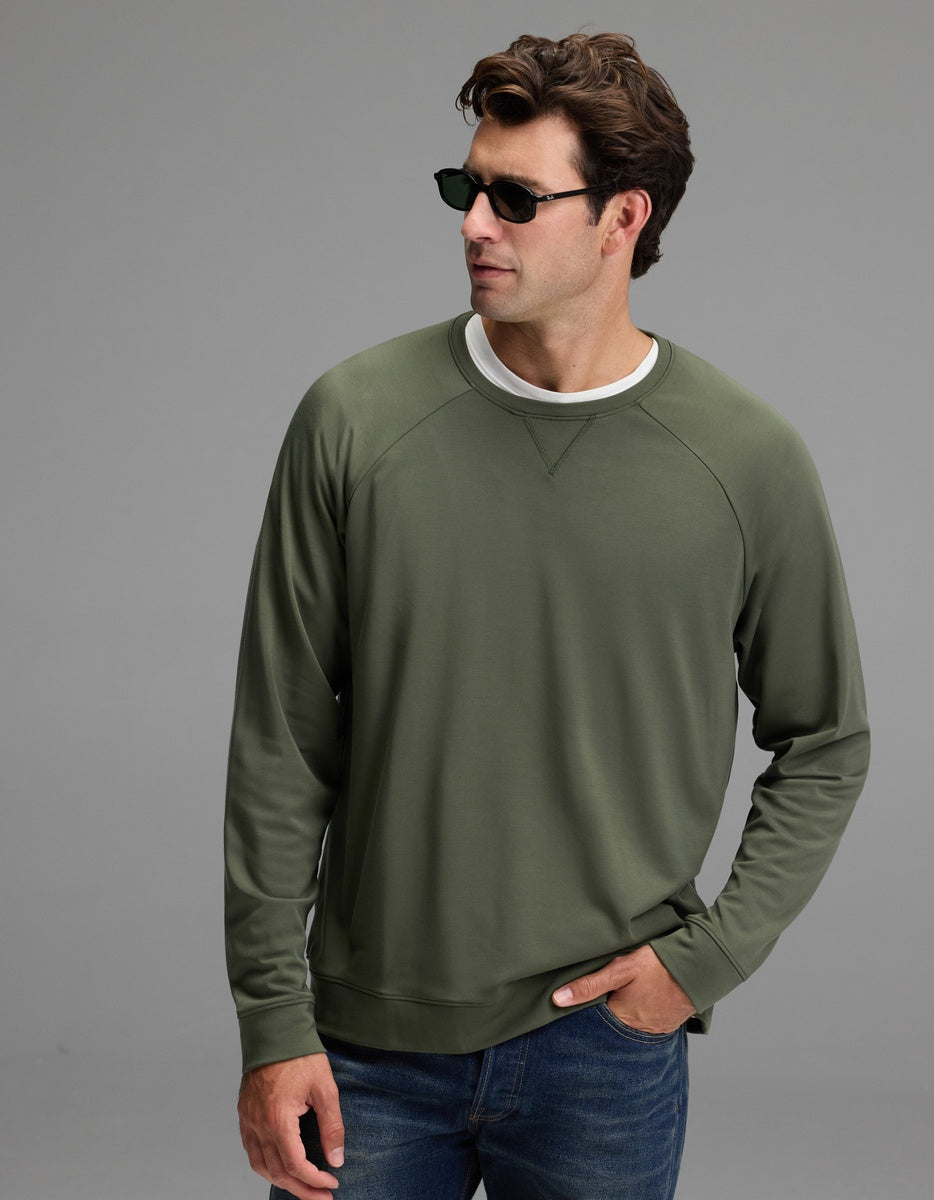 A man stands against a plain gray background with one hand in his jeans pocket, wearing dark sunglasses, a white T-shirt, and the Cozy Earth Men’s Brushed Bamboo Pullover Crew in Olive.