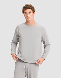 A young man with curly hair wears Cozy Earth's Men's Brushed Bamboo Pullover Crew in light gray, standing against a plain gray background and holding the hem of his sweatshirt. |Color:Stone