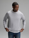 A man wearing the Cozy Earth Men’s Brushed Bamboo Pullover Crew in Stone adjusts his shirt while standing against a plain gray background.