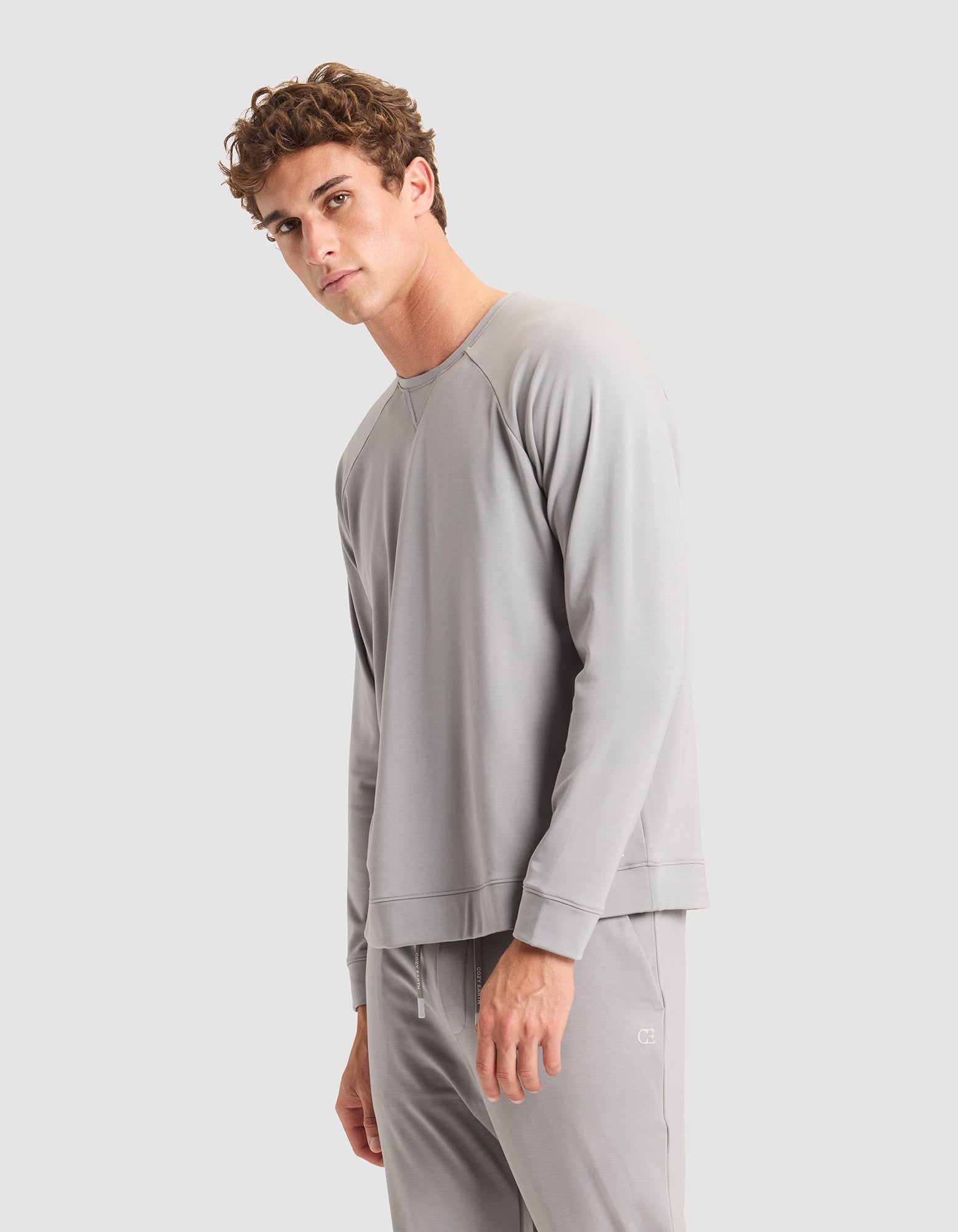 A young man with curly brown hair models the Cozy Earth Men's Brushed Bamboo Pullover Crew in light gray, paired with matching pants, standing against a plain light gray background and looking slightly toward the camera with a neutral expression. 
