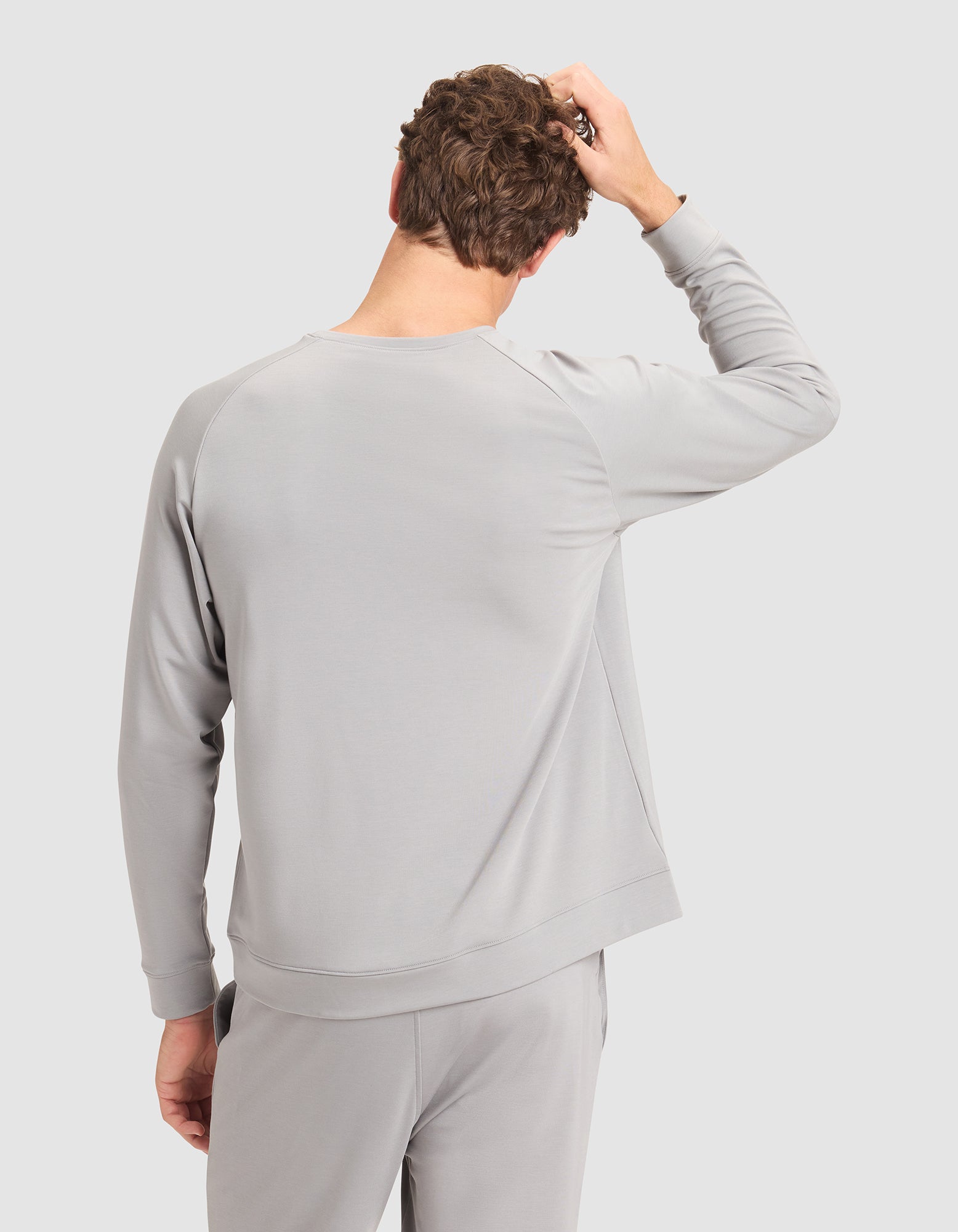 A person wearing the Cozy Earth Men’s Brushed Bamboo Pullover Crew in light gray stands with their back to the camera, right hand on their head, against a plain light background. 