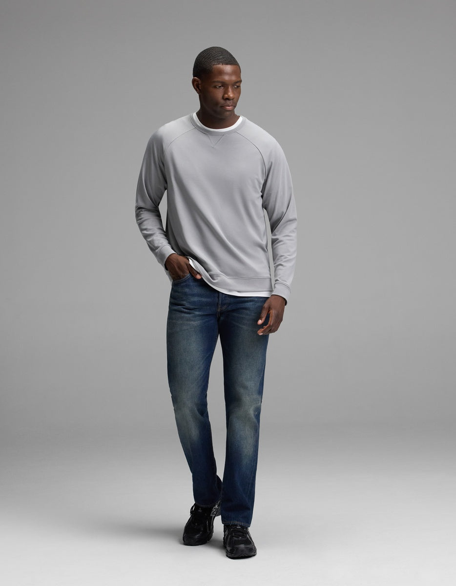 A man wearing the Cozy Earth Men’s Brushed Bamboo Pullover Crew in light gray, paired with blue jeans and black shoes, stands against a plain light gray background, looking to his right with one hand in his pocket.